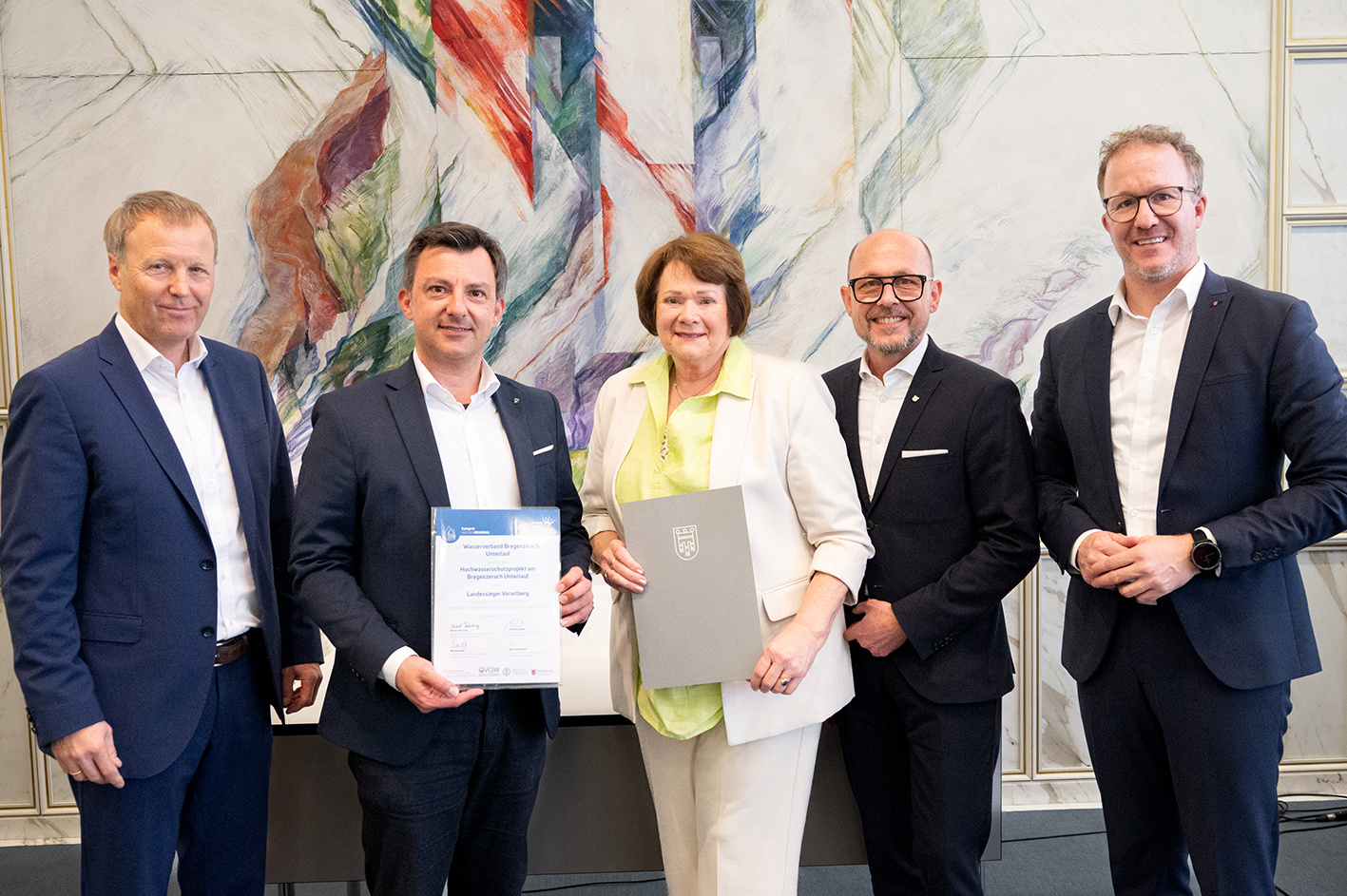 Gruppenfoto mit Bürgermeister Staudinger, einer Frau und drei Männer blicken vor einer bunten Wand in die Kamera. Er hält eine Urkunde in Händen.