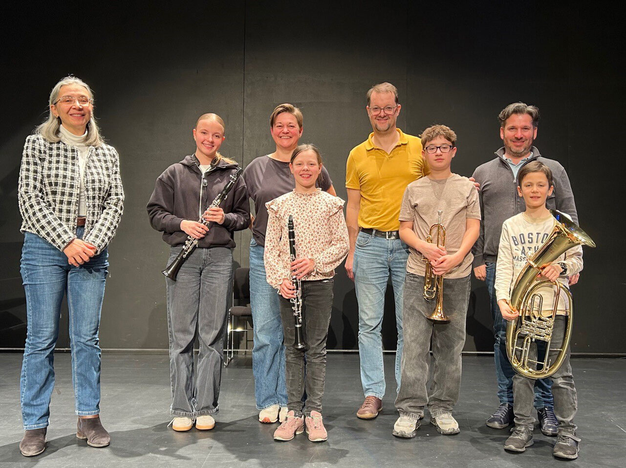Vier Kinder mit Instrumenten in der Hand und vier Erwachsene stehen vor einer grauen Wand und lächeln in die Kamera