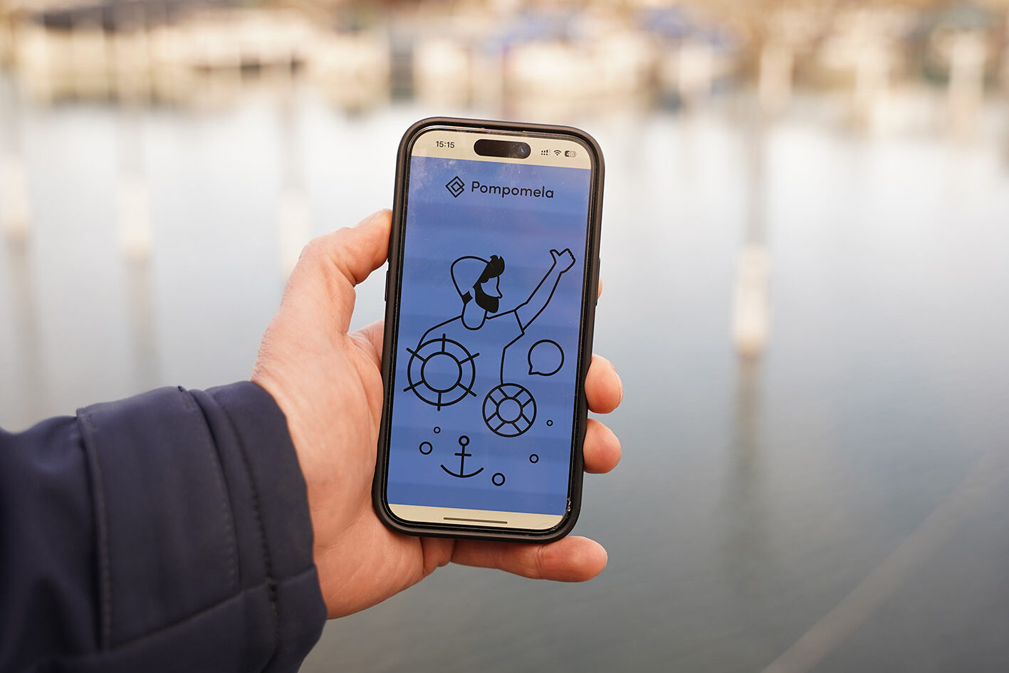 Eine Hand hält ein Smartphone auf dessen Bildschirm die Einstiegsseite der Pompomela-App zu sehen ist.