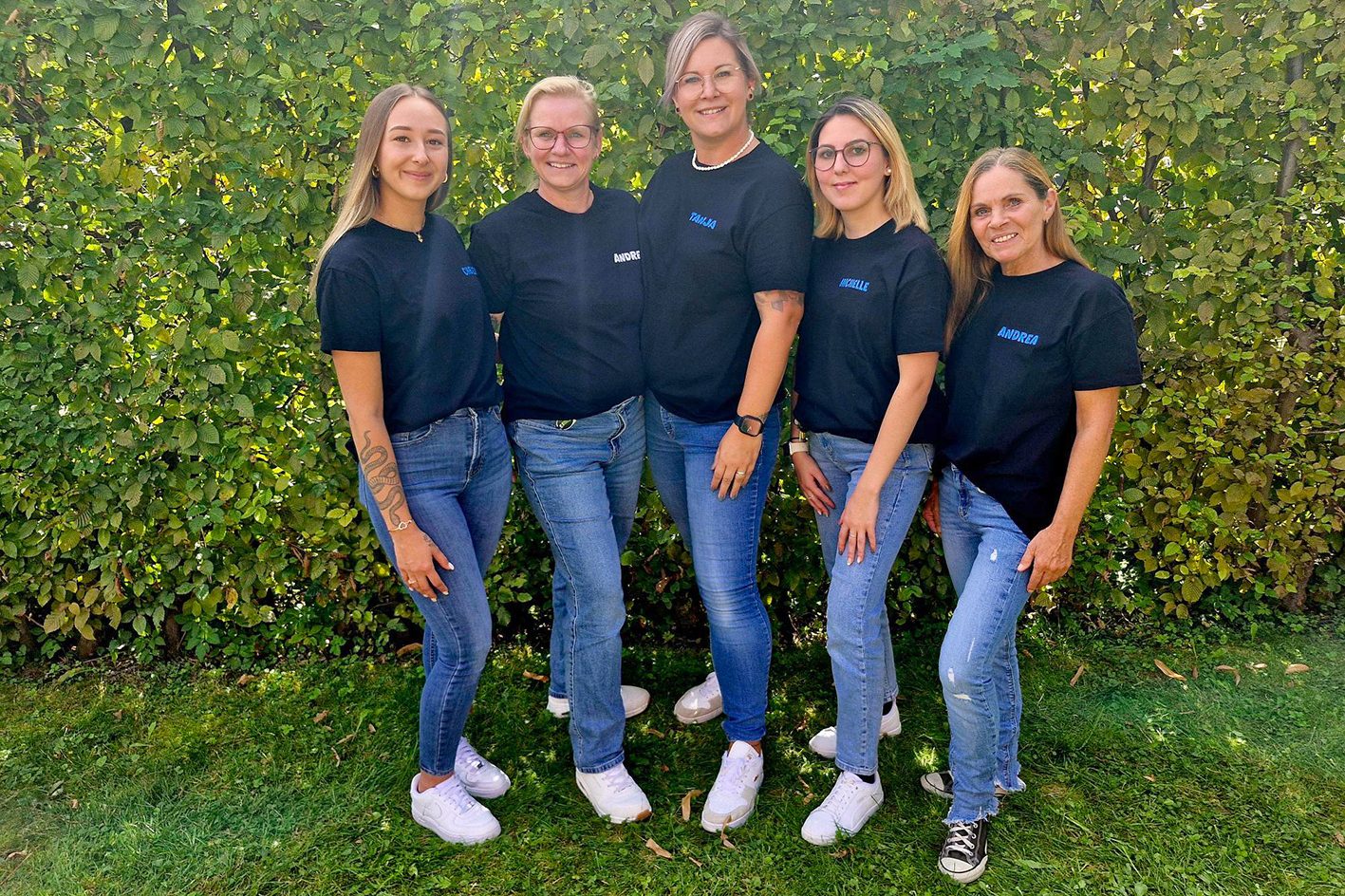 Fünf Kleinkindbetreuerinnen stehen lächelnd auf dem Rasen vor einer Hecke.