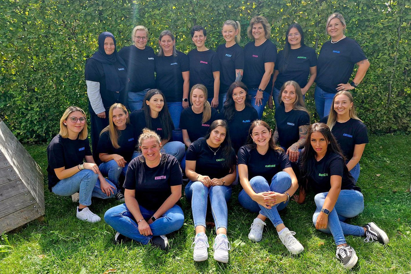 19 Kleinkindbetreuerinnen lächeln vor einer Hecke in die Kamera.