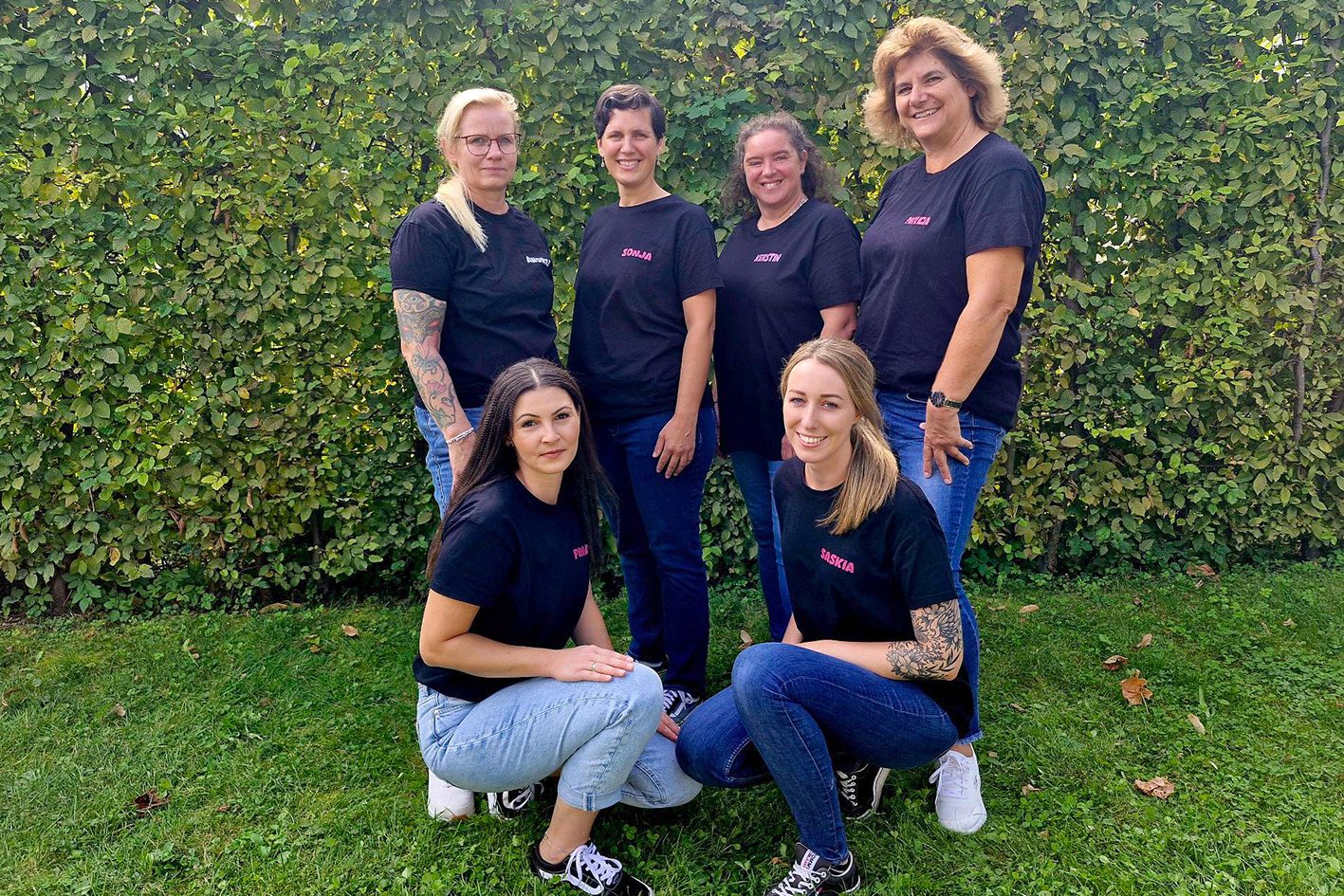 Sechs Kleinkindbetreuerinnen lächeln vor einer Hecke. Vier Personen stehen, zwei Personen knien.