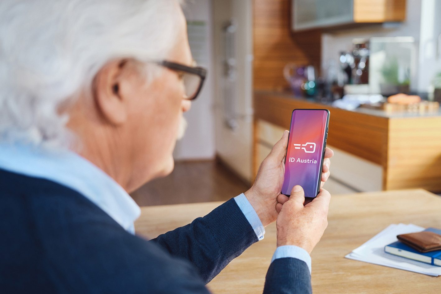 Ein grauhaariger Mann sitzt in einer Küche am Tisch und blickt auf ein Handy in seiner Hand, auf dessen Bildschirm das ID Austria Logo abgebildet ist.