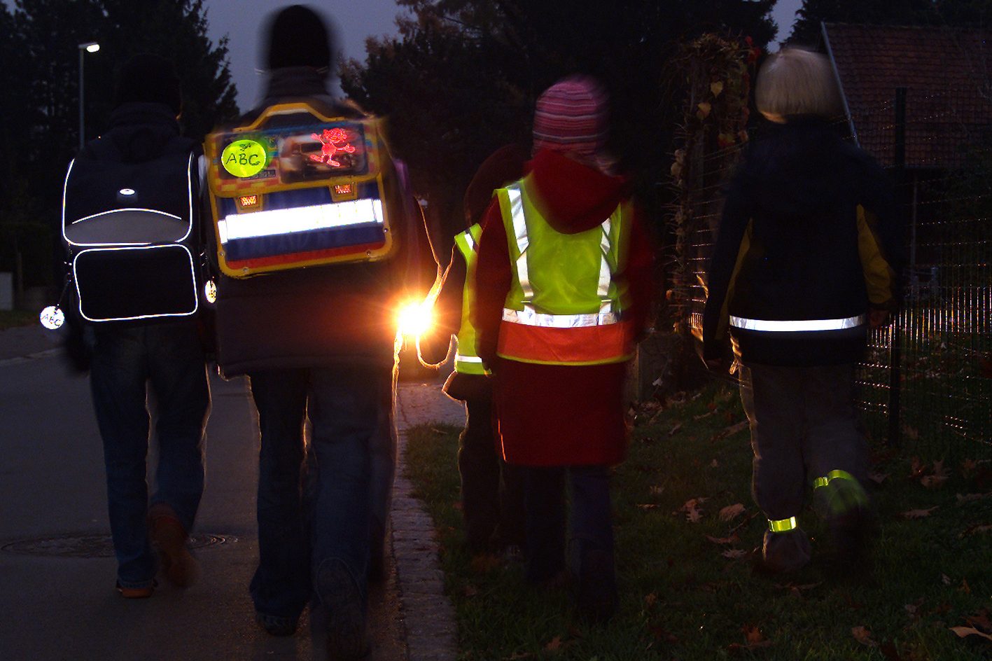 Fünf Kinder gehen in der Dunkelheit am Straßenrand entlang und sind von hinten zu sehen. Zwei tragen reflektierende Schultaschen am Rücken, zwei reflektierende Warnwesten und ein Kind eine reflektierenden Jacke