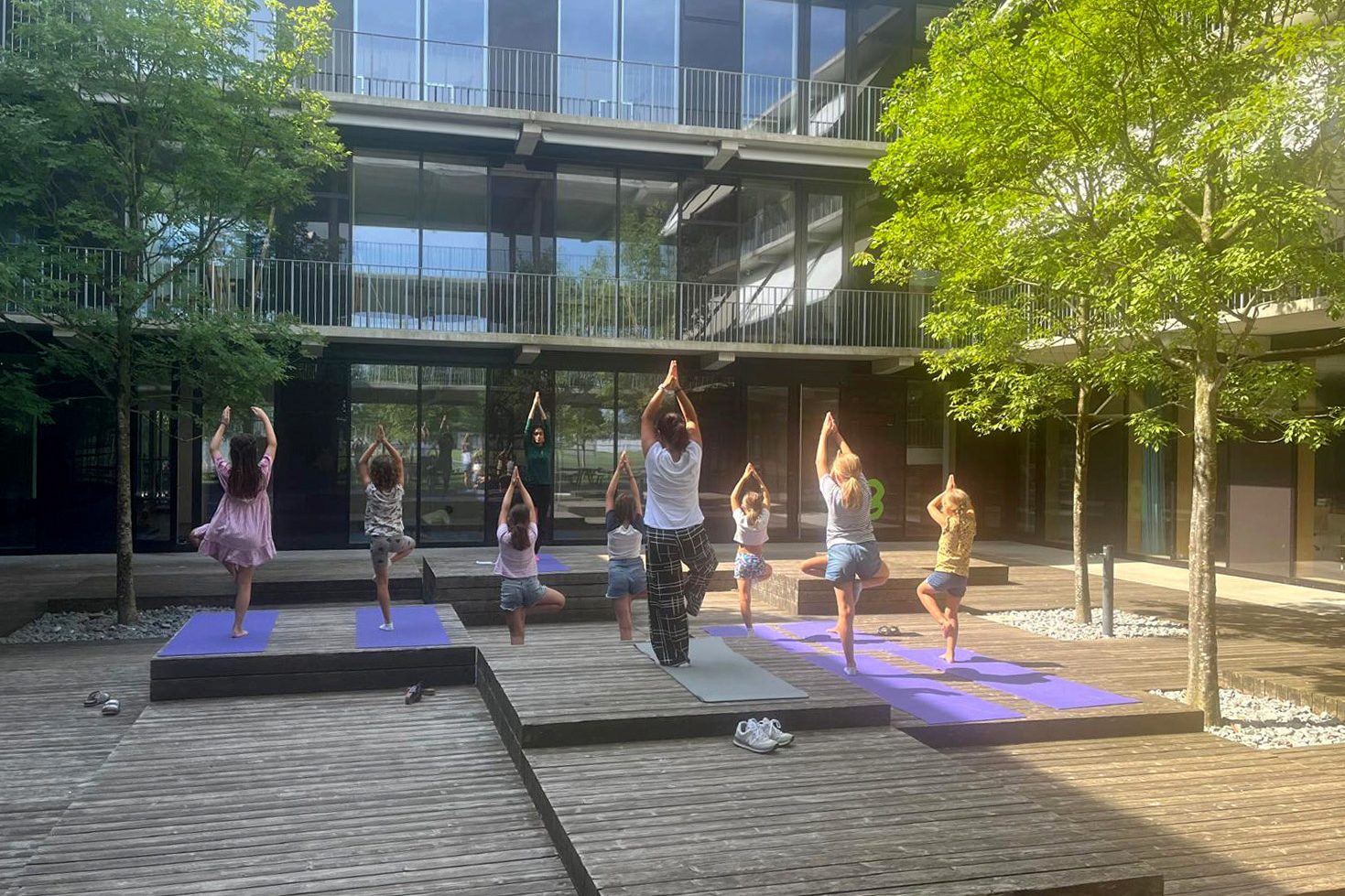 Sechs Mädchen und eine Erwachsene stehen mit dem Rücken zur Kamera und führen eine Yogaübung aus