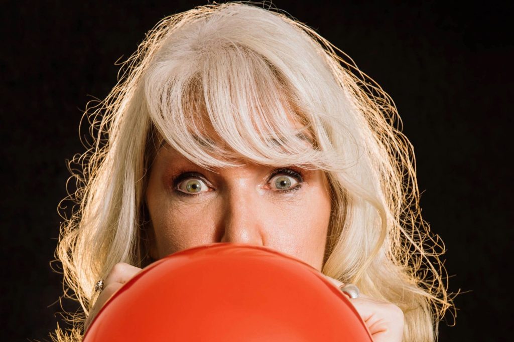 Die blonde Kabarettistin Barbara Balldini blick mit weit aufgerissenen Augen in die Kamera und pustet vor schwarzem Hintergrund einen roten Luftballon auf.