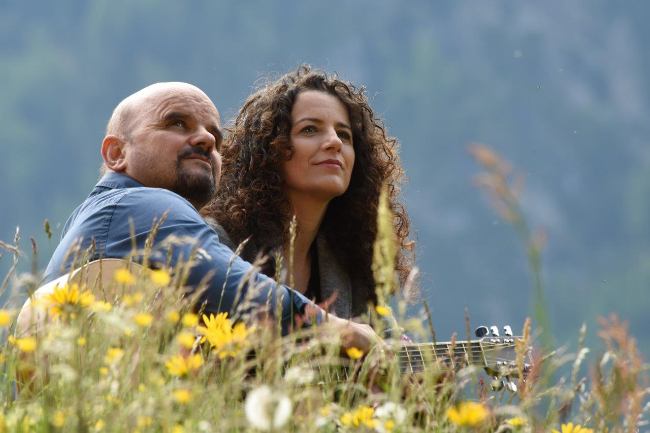 Das Musikduo Bluatschink sitzt mit einer Gitarre auf einer blühenden Blumenwiese.