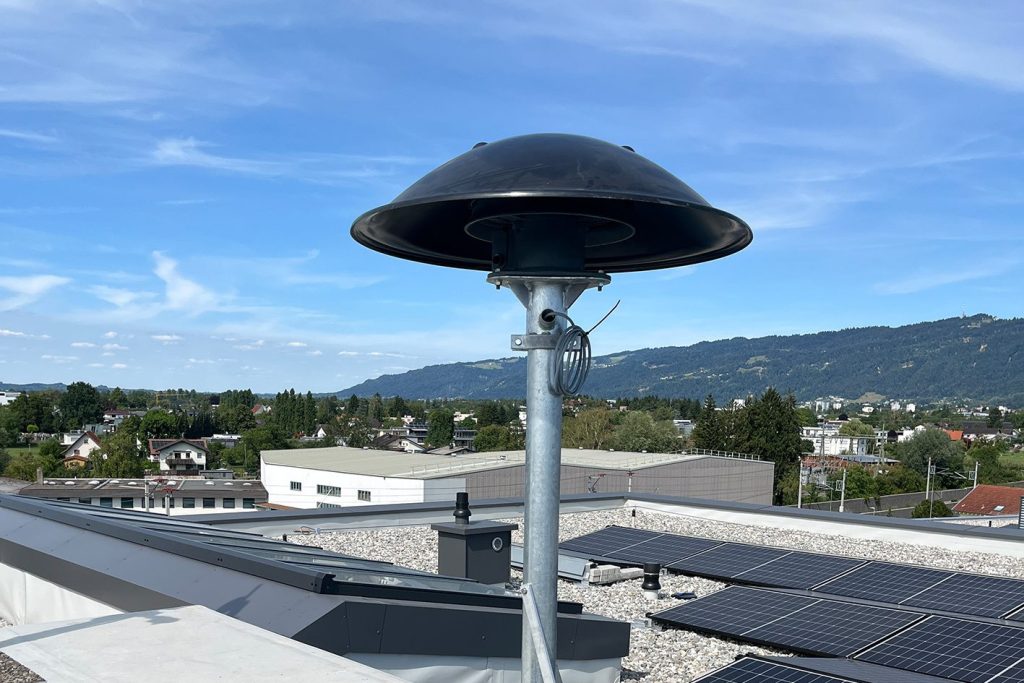 Eine Sirene auf einem Flachdach mit Solarmodulen vor einer bergigen Landschaft und blauem Himmel.