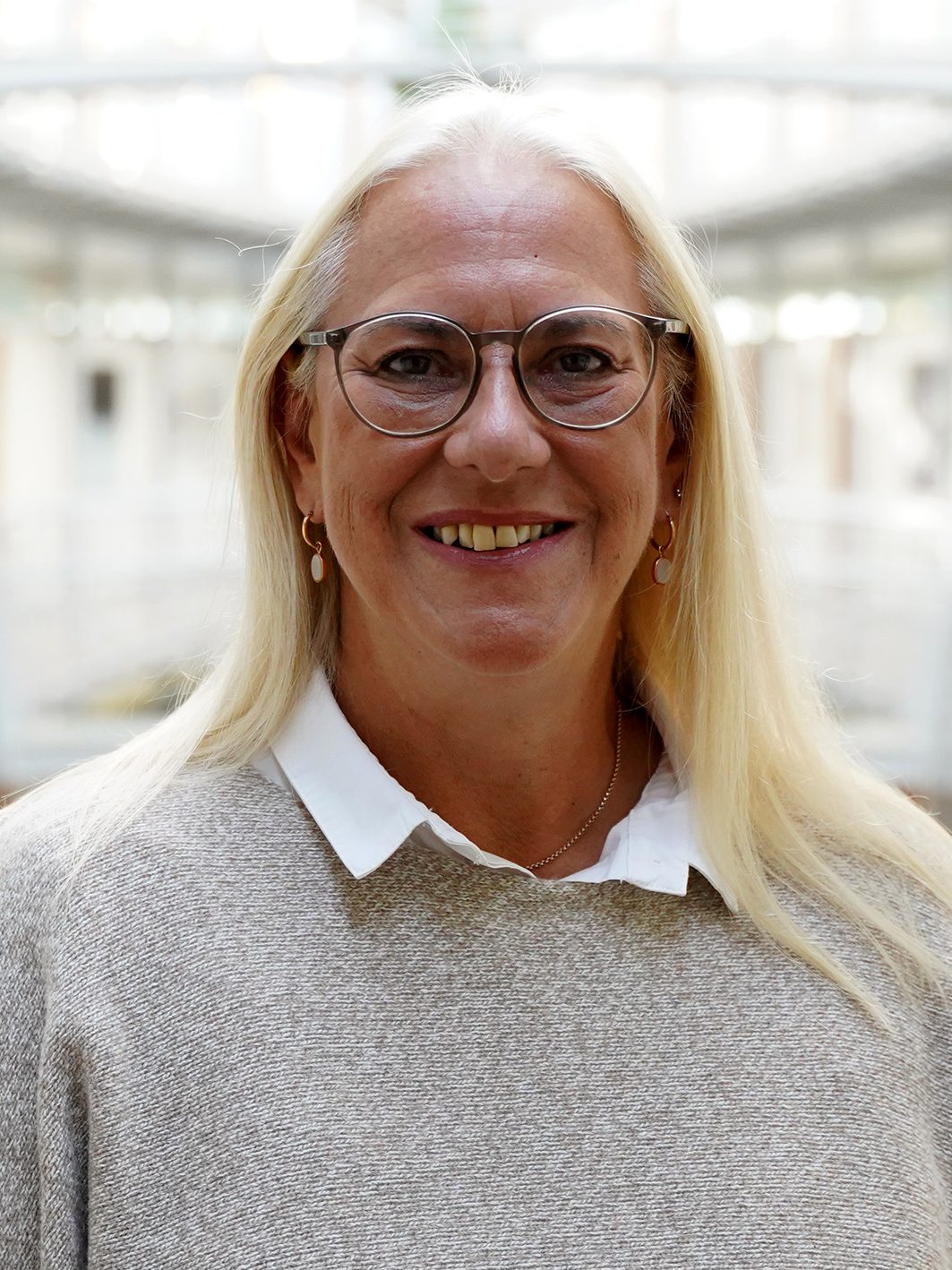 Porträt von Brigitta Schuller, sie lächelt, trägt eine Brille und hat langen blonde Haare.