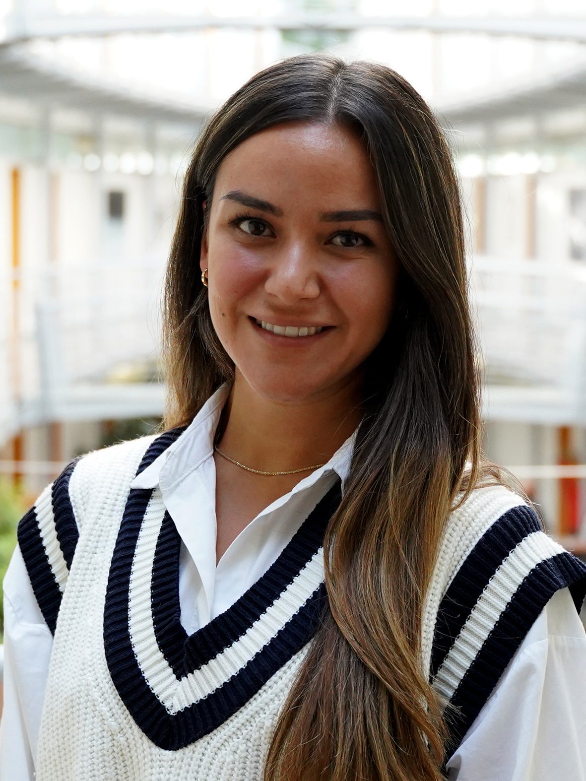 Porträt von Seda Kalkan-Deniz, sie lächelt, hat lange, dunkle Haaren und trägt einen weißen Strickpullunder mit einer weißen Bluse darunter.